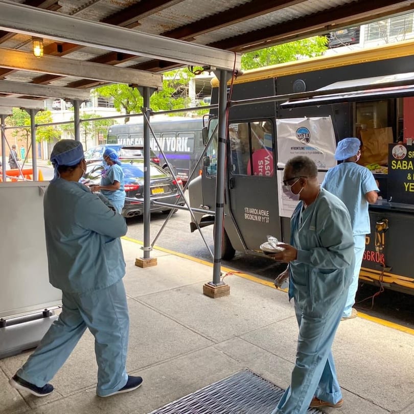 Healthcare workers in scrubs and masks receive food from a Yemen Cafe & Restaurant food truck.