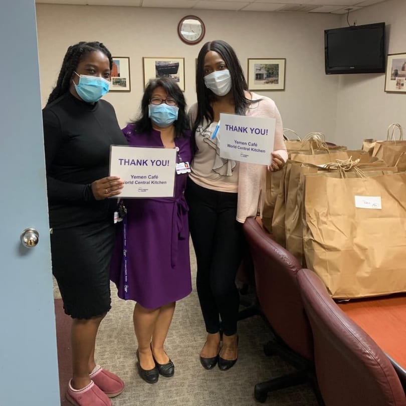 Three women in masks hold "Thank You" signs from Yemen Cafe, surrounded by many brown paper bags.