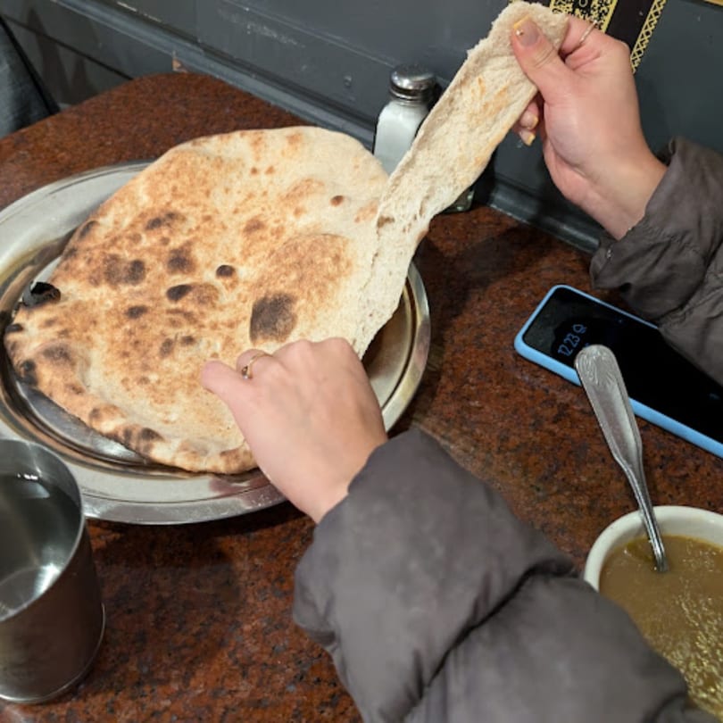 Hands tearing a piece of Yemeni flatbread from a large round on a metal platter, with a bowl of soup nearby.