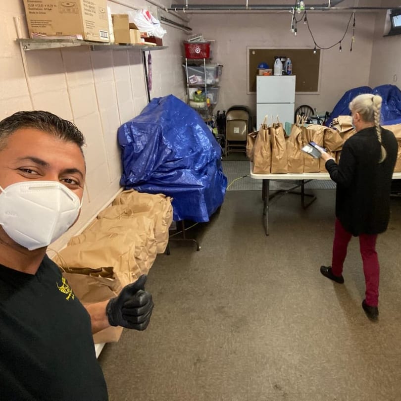 Man in a mask gives a thumbs up while a woman prepares brown paper bags of food in a storage room.