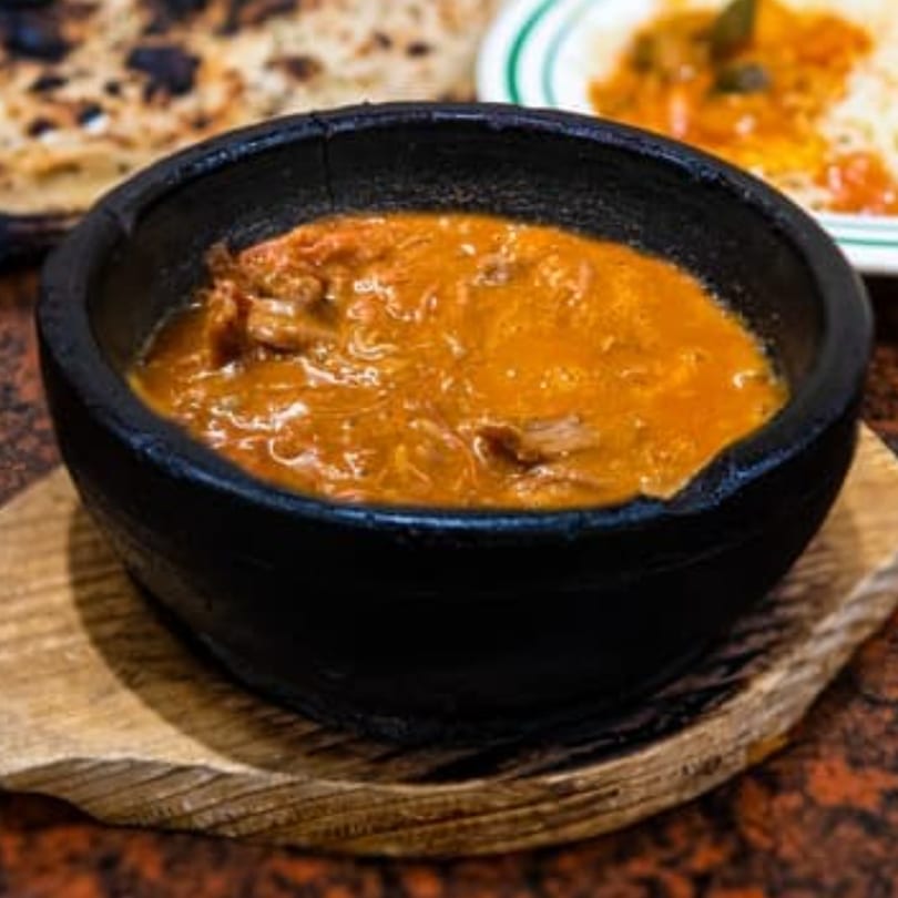 A black bowl of orange stew with meat, served on a wooden board with bread and rice.