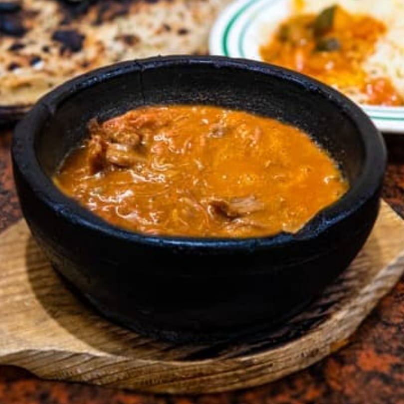 A black bowl of orange stew, possibly lamb or chicken, on a wooden board.