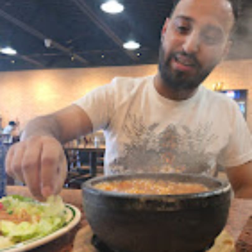 A man in a white shirt eating a steaming bowl of Middle Eastern food with a side salad.