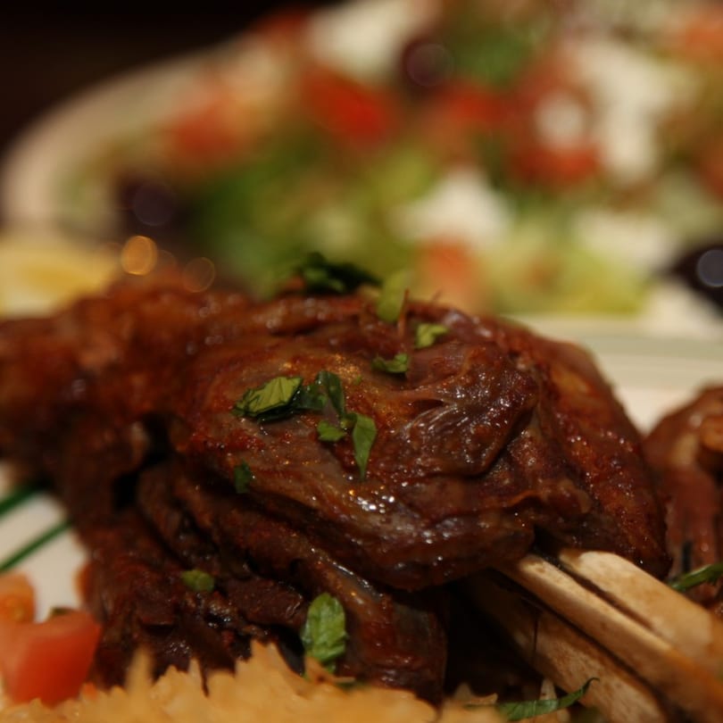 Close-up of a large, dark brown roasted lamb shank garnished with fresh green herbs, with salad in background.