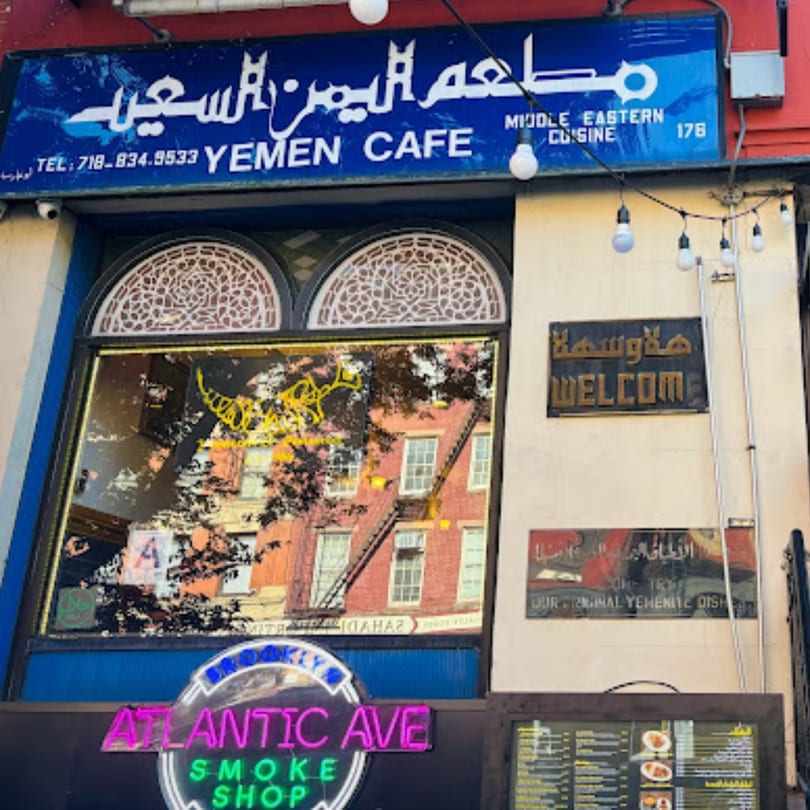 Exterior of Yemen Cafe & Restaurant with blue signage, arched windows, and a neon Atlantic Ave Smoke Shop sign.