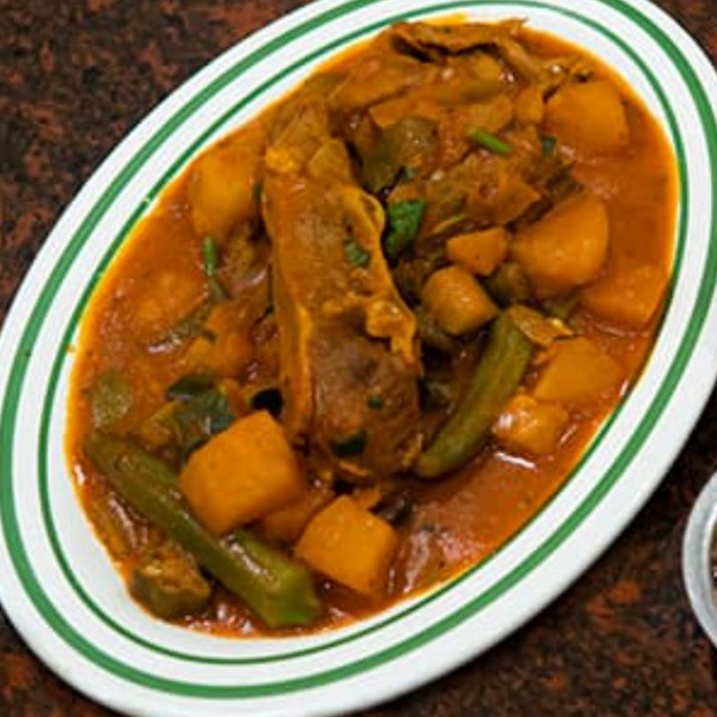A white oval plate with green stripes holds a hearty stew of meat, potatoes, and okra in a rich orange sauce.